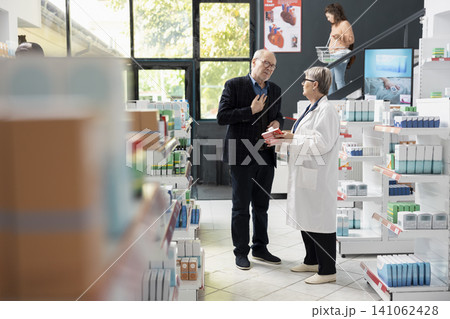 Female pharmacist helps senior customer with appropriate medication while standing near organized pharmacy shelves. Medical expert explains prescription details and offers guidance in modern drugstore Female pharmacist helps senior customer with appropriate medication while standing near organized pharmacy shelves. Medical expert explains prescription details and offers guidance in modern drugstore 141062428