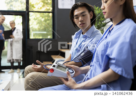 Adult woman in blue scrubs checks blood pressure asian man during routine medical visit. Healthcare professional ensures accurate screening and patient trust in clinical waiting room. Adult woman in blue scrubs checks blood pressure asian man during routine medical visit. Healthcare professional ensures accurate screening and patient trust in clinical waiting room. 141062436