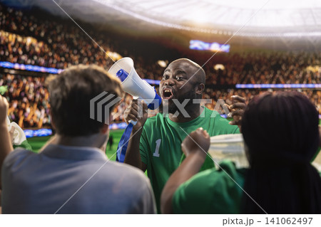 Close up of intense football supporter using megaphone to lead stadium crowd during match day. Passionate fan chants loudly from sports venue stands, driving crowd energy Close up of intense football supporter using megaphone to lead stadium crowd during match day. Passionate fan chants loudly from sports venue stands, driving crowd energy 141062497