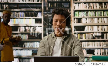 Student with headphones reading open book and studying with focus music, multitasking at desk with laptop and study materials during study session with peers. Quiet library space. Student with headphones reading open book and studying with focus music, multitasking at desk with laptop and study materials during study session with peers. Quiet library space. 141062517