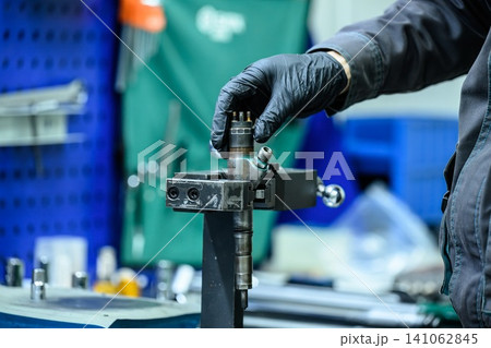 Mechanic hand in glove adjusting common rail diesel fuel injector fixed in clamp holder during repair in workshop Mechanic hand in glove adjusting common rail diesel fuel injector fixed in clamp holder during repair in workshop 141062845