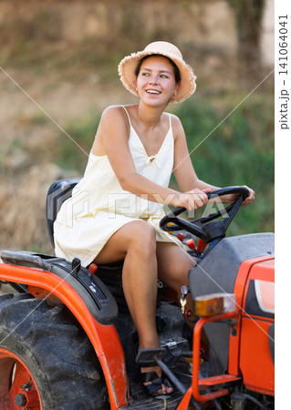 Young woman sits on minitractor and drives rural vehicle, performs loading work on farm Young woman sits on minitractor and drives rural vehicle, performs loading work on farm 141064041