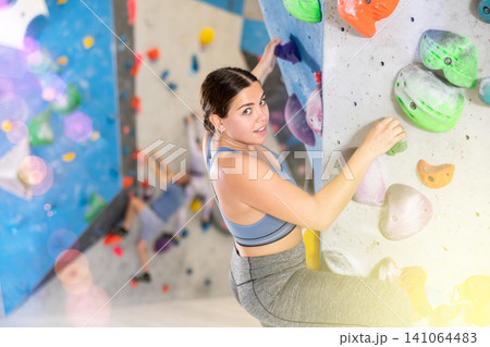 Cheerful young girl climbing artificial wall in bouldering center Cheerful young girl climbing artificial wall in bouldering center 141064483