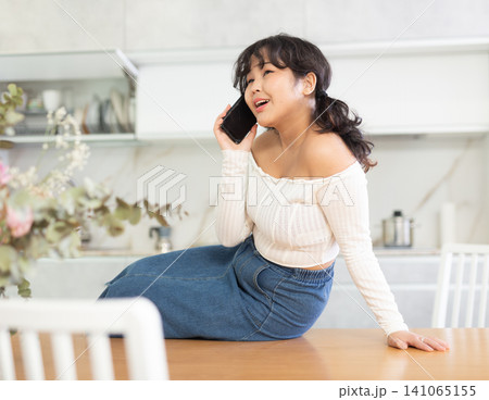 Young girl cheerfully chatting on a mobile phone while sitting on a table in home interior Young girl cheerfully chatting on a mobile phone while sitting on a table in home interior 141065155