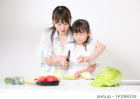 キッチンで料理を通して包丁の使い方を学ぶ母親(親子)食育を実践する母親と小学生や幼児の女の子 キッチンで料理を通して包丁の使い方を学ぶ母親(親子)食育を実践する母親と小学生や幼児の女の子 141066108
