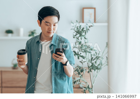 Happy Asian man drinking hot coffee in the morning using the phone while standing in the living room Happy Asian man drinking hot coffee in the morning using the phone while standing in the living room 141066997