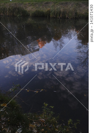 Still water reflects an old building, soft clouds, and bare tree branches 141071430