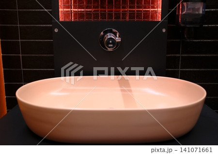 Elegant, light pink oval sink glows warmly, set against a backdrop of black tiles and a vibrant red-lit panel Elegant, light pink oval sink glows warmly, set against a backdrop of black tiles and a vibrant red-lit panel 141071661