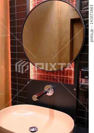 Modern restroom: round mirror, wall faucet, oval sink, warm orange grid lighting 141071663