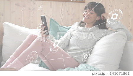 Reclining woman wearing gray sweater, pink leggings scrolling smartphone at home, with pillows 141071861