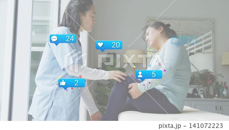 Leaning Korean woman in light-blue scrubs checking man holding knee during home visit, social icons 141072223