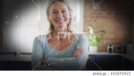 Smiling woman in blue top, thin-chain posing arms crossed in home kitchen, gold bokeh, coffee maker 141072415
