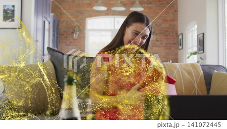 Holding red gift, woman in red top smiling in living room with laptop, champagne and glitter Holding red gift, woman in red top smiling in living room with laptop, champagne and glitter 141072445