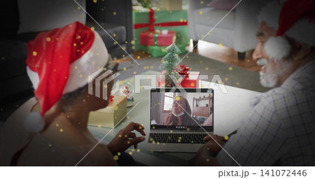 Smiling couple wearing red-and-white Santa hats waving at laptop on living room table, with gifts Smiling couple wearing red-and-white Santa hats waving at laptop on living room table, with gifts 141072446