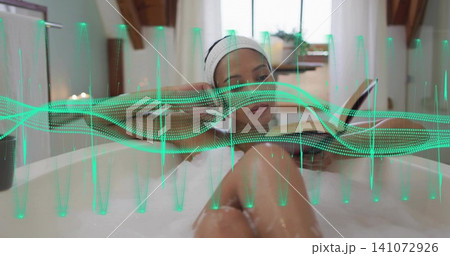 Reclining mid-adult woman reading book in home tub, with green waveform, white headband and towel Reclining mid-adult woman reading book in home tub, with green waveform, white headband and towel 141072926