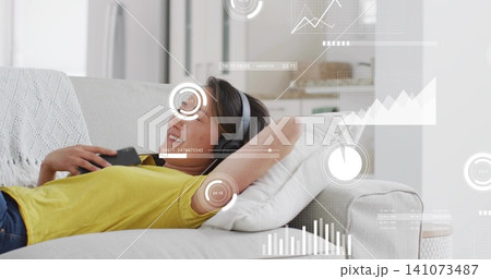 Reclining Korean woman wearing yellow top and grey headphones, holding phone on sofa with AR HUD 141073487