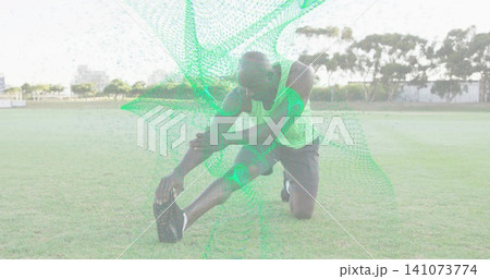 Kneeling man reaching forward touching toes on sports field, neon top dark shorts mesh overlay 141073774