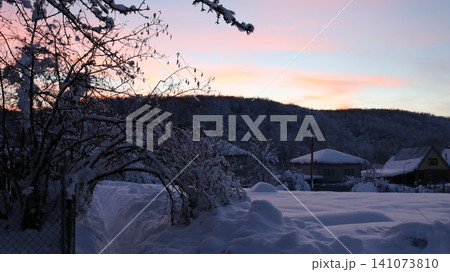 A snow-covered rural landscape. In the foreground are bushes and a path covered in thick snow, while in the distance, white-roofed houses and a wooded hill are visible against a soft pink-blue sky. A snow-covered rural landscape. In the foreground are bushes and a path covered in thick snow, while in the distance, white-roofed houses and a wooded hill are visible against a soft pink-blue sky. 141073810