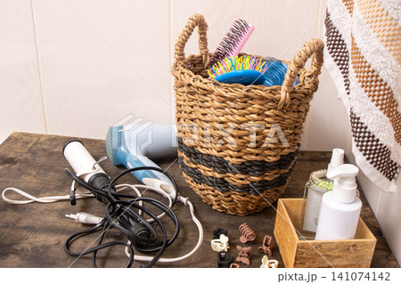 Bathroom Counter Organization with Woven Basket. Hairdryer, towel, curling iron and combs in the bathroom at home. Hairpins and hair care and styling items. Bathroom Counter Organization with Woven Basket. Hairdryer, towel, curling iron and combs in the bathroom at home. Hairpins and hair care and styling items. 141074142