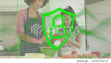 Stirring mother wearing apron, child in patterned dress helping in kitchen with green bowl, padlock 141074217