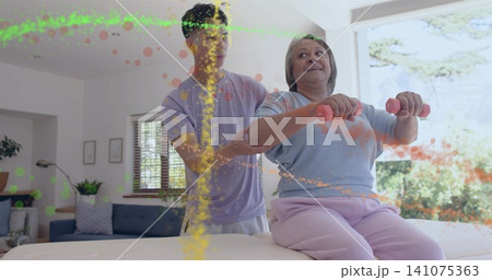 Guiding senior woman in blue top holding pink weights, caregiver supporting on padded table at home 141075363