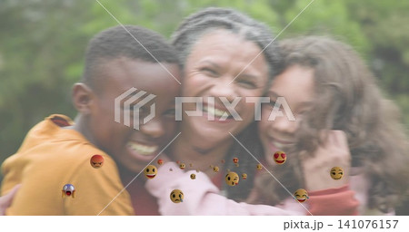 Smiling mother in red top and children leaning into hug at park, showing floating emoji reactions Smiling mother in red top and children leaning into hug at park, showing floating emoji reactions 141076157