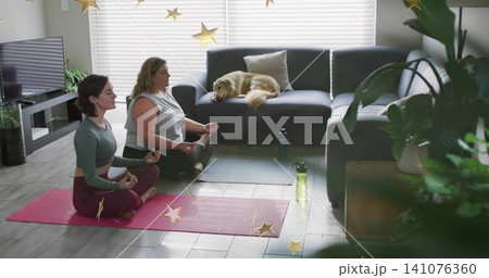 Meditating two women seated crosslegged on yoga mats at home, in activewear with golden retriever 141076360