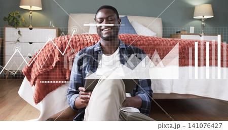 Smiling man in plaid shirt sitting on wooden bedroom floor, leaning on bed, holding tablet overlays Smiling man in plaid shirt sitting on wooden bedroom floor, leaning on bed, holding tablet overlays 141076427