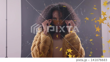 Pulling yellow aviator shades down, woman posing in studio with tan faux-fur coat, gold confetti Pulling yellow aviator shades down, woman posing in studio with tan faux-fur coat, gold confetti 141076683