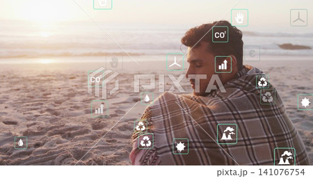 Sitting man wrapped in plaid blanket gazing toward sea at sunset, with eco icons, copy space Sitting man wrapped in plaid blanket gazing toward sea at sunset, with eco icons, copy space 141076754