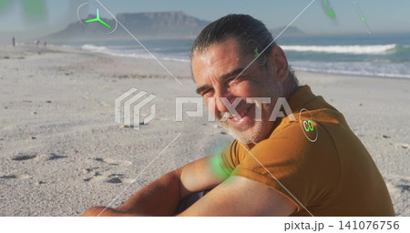 Smiling mature man wearing mustard shirt sitting on sandy beach by ocean, with green overlays 141076756