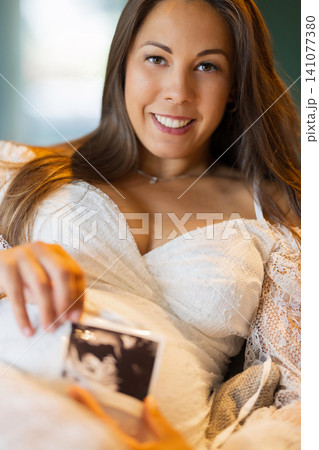 Pregnant woman smiling and holding an ultrasound photo at home 141077380