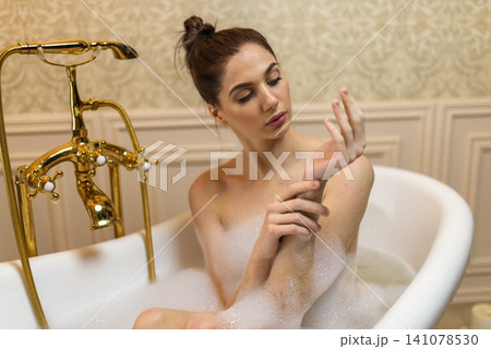 Thoughtful woman sitting in a bubble bath next to a vintage tap 141078530