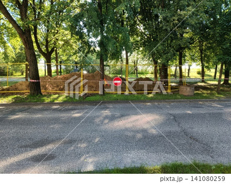 Yellow metal fence blocks access to dirt piles in park under leafy green trees. Red no entry sign attached. Sunlight filters through canopy. Calm daytime scene with natural shadows Yellow metal fence blocks access to dirt piles in park under leafy green trees. Red no entry sign attached. Sunlight filters through canopy. Calm daytime scene with natural shadows 141080259