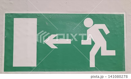 Green emergency exit sign with white running figure and upward arrow on white wall 141080278