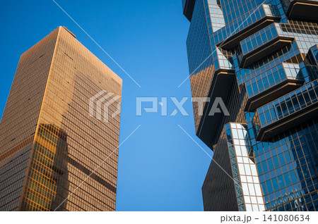 Tall golden bronze glass skyscraper stands alongside unique blue octagonal geometric office tower reaching toward clear blue daytime sky showing tiny white moon Tall golden bronze glass skyscraper stands alongside unique blue octagonal geometric office tower reaching toward clear blue daytime sky showing tiny white moon 141080634