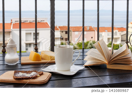 Breakfast table on a balcony in Makarska, Croatia, with coffee cup, open book, bread with butter, and Adriatic Sea view Breakfast table on a balcony in Makarska, Croatia, with coffee cup, open book, bread with butter, and Adriatic Sea view 141080987