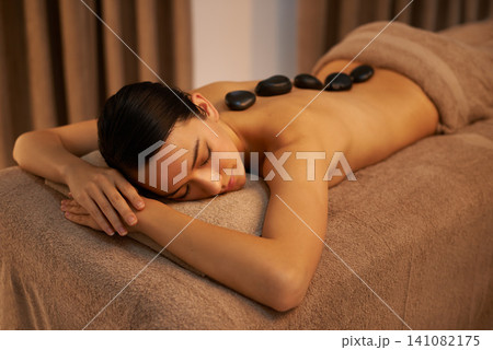 Bed, woman and massage with stone at spa for stress relief, organic body health and detox. Towel, relax and zen female person with hot rock for wellness, self care and spiritual healing at resort Bed, woman and massage with stone at spa for stress relief, organic body health and detox. Towel, relax and zen female person with hot rock for wellness, self care and spiritual healing at resort 141082175