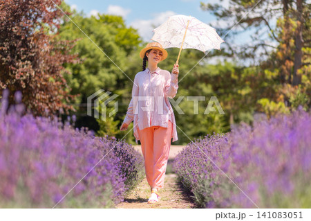 Elegant young Caucasian woman poses in blooming lavender fields in Provence, holds umbrella. Summer vacation concept with French countryside aesthetics, calm luxury, and natural beauty. Elegant young Caucasian woman poses in blooming lavender fields in Provence, holds umbrella. Summer vacation concept with French countryside aesthetics, calm luxury, and natural beauty. 141083051