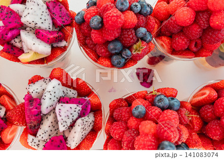 Plastic takeaway cups packed with colorful fresh fruit are offered at a busy outdoor fruit stall. Summer market atmosphere celebrates seasonal harvest, nutrition, and convenient snacks. Plastic takeaway cups packed with colorful fresh fruit are offered at a busy outdoor fruit stall. Summer market atmosphere celebrates seasonal harvest, nutrition, and convenient snacks. 141083074