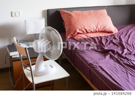 A white fan is positioned on a chair next to a bedside table and a rumpled bed with purple sheets and an orange pillow. A white fan is positioned on a chair next to a bedside table and a rumpled bed with purple sheets and an orange pillow. 141084266