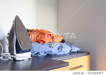 An electric iron sits next to a pile of folded laundry on a dark surface. An electric iron sits next to a pile of folded laundry on a dark surface. 141084267