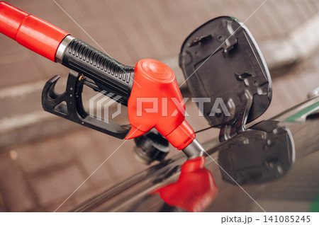 Red fuel nozzle inserted into a car gas tank at a petrol station close-up, fuel refilling and transport economy concept Red fuel nozzle inserted into a car gas tank at a petrol station close-up, fuel refilling and transport economy concept 141085245