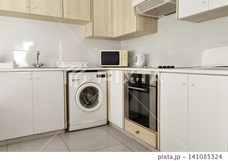 Modern compact kitchen interior with built-in appliances, washing machine, microwave, electric kettle and white cabinets. Bright functional home cooking space with contemporary design. Modern compact kitchen interior with built-in appliances, washing machine, microwave, electric kettle and white cabinets. Bright functional home cooking space with contemporary design. 141085324