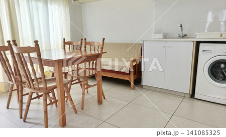 Bright open-plan apartment dining area with compact kitchenette, wooden table and chairs, sofa, washing machine and natural daylight. Cozy furnished vacation rental interior. Bright open-plan apartment dining area with compact kitchenette, wooden table and chairs, sofa, washing machine and natural daylight. Cozy furnished vacation rental interior. 141085325