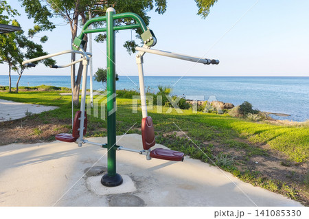Outdoor fitness equipment overlooking the sea in a coastal park. Public exercise machine by the waterfront surrounded by green trees, offering a scenic place for healthy recreation. Outdoor fitness equipment overlooking the sea in a coastal park. Public exercise machine by the waterfront surrounded by green trees, offering a scenic place for healthy recreation. 141085330