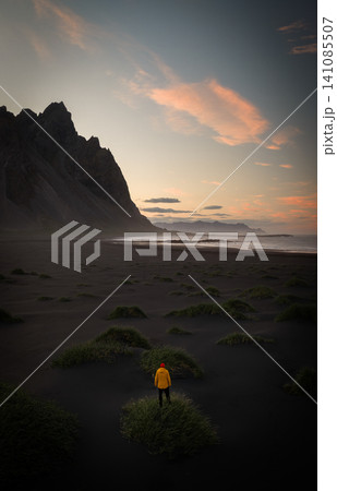 Lone traveler in a yellow jacket standing on the black sand beach of Vestrahorn at sunset. Dramatic mountains, soft evening light and Icelandic coastline create a breathtaking landscape. 141085507