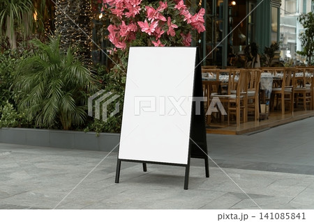 Blank white outdoor advertising stand sandwich board mock up template. Clear street signage board placed by an outdoor dinning area of a restaurant. 141085841