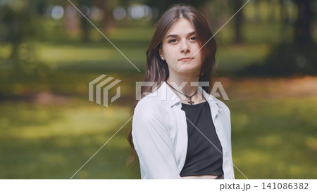Confident woman with flowing brown hair standing in urban green space, wearing white shirt and black top, embodying urban lifestyle Confident woman with flowing brown hair standing in urban green space, wearing white shirt and black top, embodying urban lifestyle 141086382
