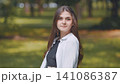 Young woman with long brown hair wearing a white shirt and black top standing in a park with blurred background 141086387